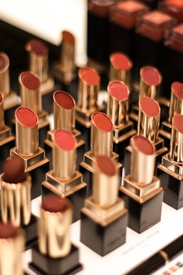 A display of various lipsticks, each housed in a gold and black casing, arranged neatly in rows. The lipsticks come in different shades of red and brown, and the background is slightly blurred, emphasizing the lipsticks.