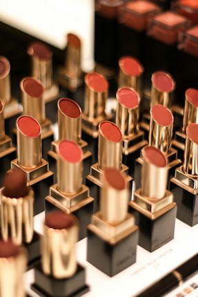 A display of various lipsticks, each housed in a gold and black casing, arranged neatly in rows. The lipsticks come in different shades of red and brown, and the background is slightly blurred, emphasizing the lipsticks.