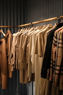 A collection of neatly arranged beige and brown garments hanging on wooden hangers. The clothing includes coats, jackets, and shirts, showcasing a range of textures such as quilted, smooth, and plaid patterns. The setting appears to be a retail space with a linear, structured display.