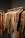 A collection of neatly arranged beige and brown garments hanging on wooden hangers. The clothing includes coats, jackets, and shirts, showcasing a range of textures such as quilted, smooth, and plaid patterns. The setting appears to be a retail space with a linear, structured display.