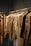 A collection of neatly arranged beige and brown garments hanging on wooden hangers. The clothing includes coats, jackets, and shirts, showcasing a range of textures such as quilted, smooth, and plaid patterns. The setting appears to be a retail space with a linear, structured display.
