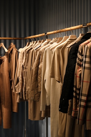 A collection of neatly arranged beige and brown garments hanging on wooden hangers. The clothing includes coats, jackets, and shirts, showcasing a range of textures such as quilted, smooth, and plaid patterns. The setting appears to be a retail space with a linear, structured display.