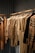 A collection of neatly arranged beige and brown garments hanging on wooden hangers. The clothing includes coats, jackets, and shirts, showcasing a range of textures such as quilted, smooth, and plaid patterns. The setting appears to be a retail space with a linear, structured display.