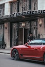 The entrance of Claridge's hotel with a doorman standing near the door. A vibrant red sports car is parked on the street in front of the hotel. The exterior features elegant wrought iron details and tall potted plants with a canopy above the entrance.