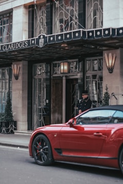 The entrance of Claridge's hotel with a doorman standing near the door. A vibrant red sports car is parked on the street in front of the hotel. The exterior features elegant wrought iron details and tall potted plants with a canopy above the entrance.