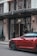 The entrance of Claridge's hotel with a doorman standing near the door. A vibrant red sports car is parked on the street in front of the hotel. The exterior features elegant wrought iron details and tall potted plants with a canopy above the entrance.