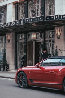 The entrance of Claridge's hotel with a doorman standing near the door. A vibrant red sports car is parked on the street in front of the hotel. The exterior features elegant wrought iron details and tall potted plants with a canopy above the entrance.
