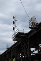 A railway signal structure is prominently featured, composed of multiple lights attached to a tall metal framework. The structure is elevated above a weathered railway bridge with crisscrossing beams. The sky overhead is overcast with thick, grey clouds casting a dull light on the scene.
