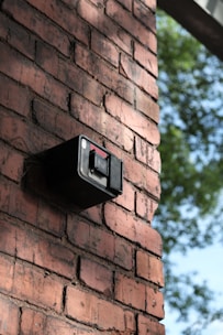 A brick wall features a mounted black security camera. The wall is in focus and displays a classic red-brown brick pattern. There is greenery visible in the background, indicating an outdoor setting.