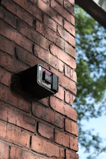 A brick wall features a mounted black security camera. The wall is in focus and displays a classic red-brown brick pattern. There is greenery visible in the background, indicating an outdoor setting.