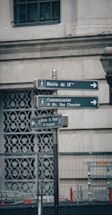 The image features a street signpost with three directional signs, located in an urban setting. The background shows part of a building wall with decorative patterns and a window. There is also a metal fence below the signs.