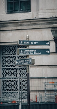 The image features a street signpost with three directional signs, located in an urban setting. The background shows part of a building wall with decorative patterns and a window. There is also a metal fence below the signs.