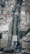 Aerial view of an urban cityscape featuring a straight, elevated railway running through the middle of densely packed buildings. The railway is flanked by roads with several vehicles, and trees line parts of the streets. The architecture includes a mix of mid-rise buildings with varied roof styles.