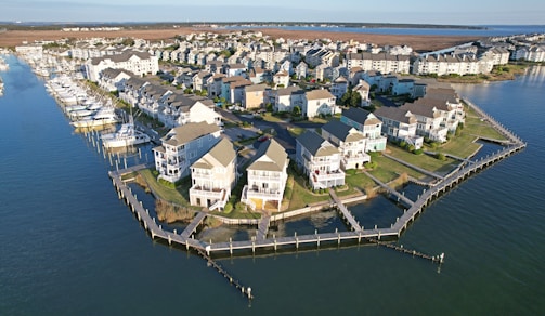 Aerial view of a coastal community with rows of houses built alongside a marina. Several boats are docked in the water area to the left, while the land is mostly occupied by closely packed residential buildings with well-maintained lawns and pathways. The scene is bordered by a large body of water, possibly a lake or the sea.