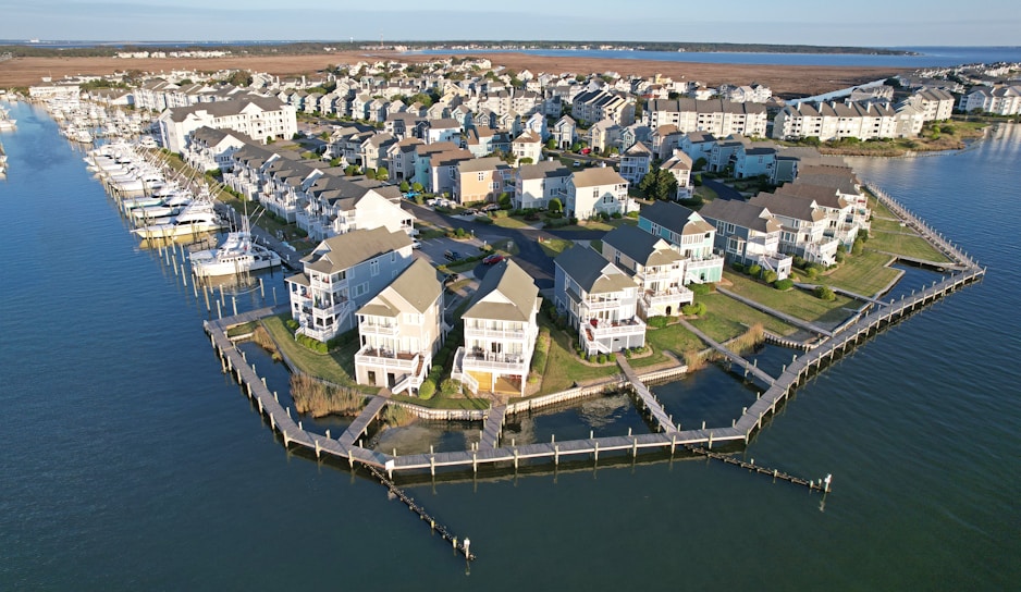 Aerial view of a coastal community with rows of houses built alongside a marina. Several boats are docked in the water area to the left, while the land is mostly occupied by closely packed residential buildings with well-maintained lawns and pathways. The scene is bordered by a large body of water, possibly a lake or the sea.