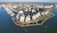 Aerial view of a coastal community with rows of houses built alongside a marina. Several boats are docked in the water area to the left, while the land is mostly occupied by closely packed residential buildings with well-maintained lawns and pathways. The scene is bordered by a large body of water, possibly a lake or the sea.