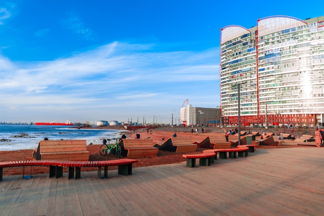 A waterfront area with modern architecture featuring a large, multi-story glass building alongside a boardwalk. There are several wooden benches and seating areas on the boardwalk. People are sitting and walking along the area. In the background, there's a large ship on the blue water, and several industrial structures are visible on the shoreline.