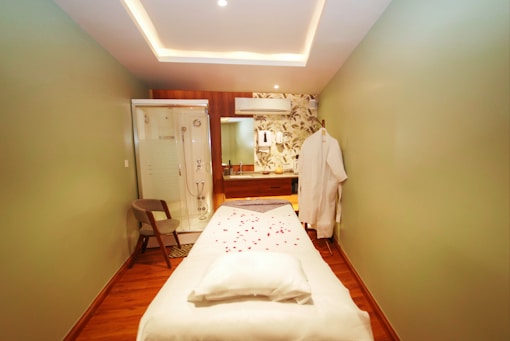 A serene and well-lit massage therapy room features a massage table adorned with scattered rose petals. The room has soft green walls with a floral accent wall in the back. A hanging white robe and a modern chair are present, along with a glass shower enclosure. The ceiling has recessed lighting, contributing to a calming atmosphere.