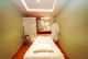 A serene and well-lit massage therapy room features a massage table adorned with scattered rose petals. The room has soft green walls with a floral accent wall in the back. A hanging white robe and a modern chair are present, along with a glass shower enclosure. The ceiling has recessed lighting, contributing to a calming atmosphere.