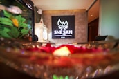 A serene spa environment featuring a prominently displayed sign with the word 'SNESAN' and a decorative emblem above it. The background has a textured wall, and to the left, there's a white lattice lantern. In the foreground, blurred rose petals and water create a tranquil and inviting ambiance.