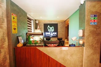 A reception area with a modern, earthy design features a wooden desk front, an aquarium with plants, and a prominent SNESAN logo in the background. The walls display multiple safety signs and instructions, and there's a small lamp and decorative items on the desk.