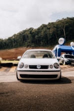 A white Volkswagen car is parked on a paved surface in an outdoor setting. There are trees and a hill in the background, suggesting a natural environment. On the right side, there is a blue inflatable structure with branding, indicating it might be part of an event or celebration. Several other parked cars are visible in the backdrop.