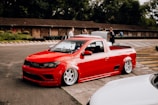 A modified red pickup truck with a sleek, lowered design is parked on a race track. The vehicle features large, polished wheels and is surrounded by a few people in the background. The setting includes trees and a rustic, wood-paneled building.