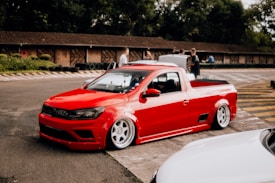 A modified red pickup truck with a sleek, lowered design is parked on a race track. The vehicle features large, polished wheels and is surrounded by a few people in the background. The setting includes trees and a rustic, wood-paneled building.