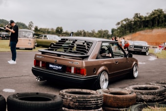 A vintage Ford Escort car with a brown exterior is prominently featured in a parking area surrounded by multiple other classic vehicles. The scene includes a photographer on the left side capturing images with a camera. Worn-out tires are placed in the foreground, and there is greenery and a dirt mound in the background.
