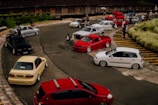 A group of modified cars is parked on a winding track, surrounded by a few people socializing. The scene is set in an open area with some vegetation and a building in the background. The cars are different colors including red, white, black, and beige.