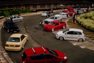 A group of modified cars is parked on a winding track, surrounded by a few people socializing. The scene is set in an open area with some vegetation and a building in the background. The cars are different colors including red, white, black, and beige.