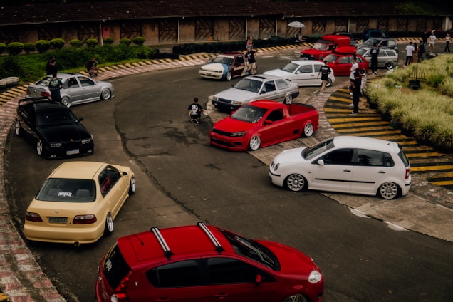 A group of modified cars is parked on a winding track, surrounded by a few people socializing. The scene is set in an open area with some vegetation and a building in the background. The cars are different colors including red, white, black, and beige.