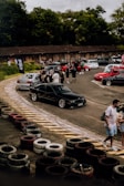 A group of people gathers around a collection of cars in an outdoor area, surrounded by greenery and a rustic wooden building. Several cars of different colors, including black, red, and white, are parked on a paved surface. The scene suggests a casual social or automotive event with participants interacting and walking around.