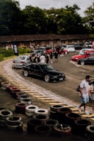 A group of people gathers around a collection of cars in an outdoor area, surrounded by greenery and a rustic wooden building. Several cars of different colors, including black, red, and white, are parked on a paved surface. The scene suggests a casual social or automotive event with participants interacting and walking around.