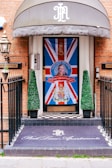 An entrance decorated with a large Union Jack flag featuring a portrait and a message 'God Save The King' in bold letters at the center. There are two conical topiary plants flanking the door. The floor has a black and white checkered pattern, and a mat at the entrance reads 'Park Lane Apartments' in elegant script. The building has a brick facade, and a metal lantern is mounted on the wall.