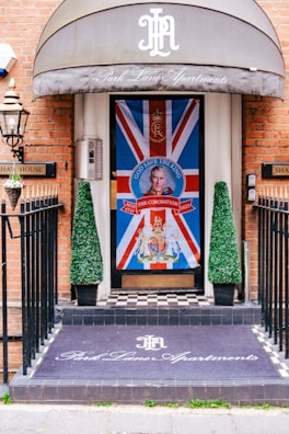 An entrance decorated with a large Union Jack flag featuring a portrait and a message 'God Save The King' in bold letters at the center. There are two conical topiary plants flanking the door. The floor has a black and white checkered pattern, and a mat at the entrance reads 'Park Lane Apartments' in elegant script. The building has a brick facade, and a metal lantern is mounted on the wall.