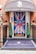 An entrance decorated with a large Union Jack flag featuring a portrait and a message 'God Save The King' in bold letters at the center. There are two conical topiary plants flanking the door. The floor has a black and white checkered pattern, and a mat at the entrance reads 'Park Lane Apartments' in elegant script. The building has a brick facade, and a metal lantern is mounted on the wall.