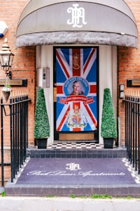 An entrance decorated with a large Union Jack flag featuring a portrait and a message 'God Save The King' in bold letters at the center. There are two conical topiary plants flanking the door. The floor has a black and white checkered pattern, and a mat at the entrance reads 'Park Lane Apartments' in elegant script. The building has a brick facade, and a metal lantern is mounted on the wall.
