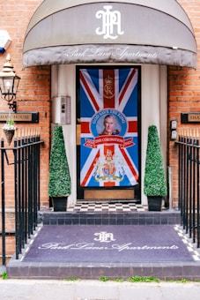 An entrance decorated with a large Union Jack flag featuring a portrait and a message 'God Save The King' in bold letters at the center. There are two conical topiary plants flanking the door. The floor has a black and white checkered pattern, and a mat at the entrance reads 'Park Lane Apartments' in elegant script. The building has a brick facade, and a metal lantern is mounted on the wall.