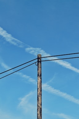 A wooden utility pole stands tall against a clear blue sky, with contrails crisscrossing in the background. Two power lines are attached to the pole, stretching out horizontally across the image.