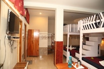 A hostel room with bunk beds and simple furniture. The room features two sets of bunk beds with white frames and dark bedding. There is a wardrobe made of wood and a flat-screen television mounted on the wall. A small table holds cups and a water bottle, and the walls are decorated with tiles and curtains, creating a warm, welcoming space.