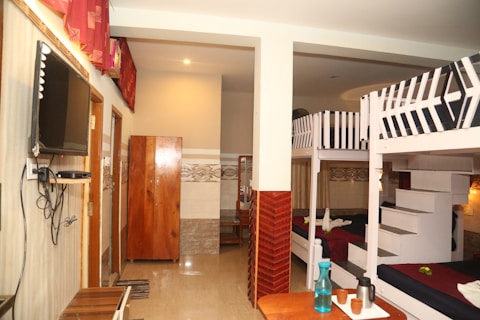 A hostel room with bunk beds and simple furniture. The room features two sets of bunk beds with white frames and dark bedding. There is a wardrobe made of wood and a flat-screen television mounted on the wall. A small table holds cups and a water bottle, and the walls are decorated with tiles and curtains, creating a warm, welcoming space.