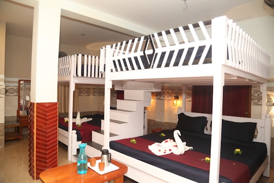 The room features a pair of white bunk beds with dark bedding. On the bottom bed, there are towels arranged in the shape of swans, alongside some small yellow flowers. A wooden tabletop in the foreground holds a blue water bottle, clay cups, and a thermos. The walls have a warm, earthy tone, accented with patterned tiles and soft lighting fixtures.
