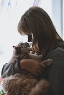 A person with medium-length hair gently holding a fluffy cat close, creating a tender moment. Soft light filters through a window in the background, illuminating the scene with a warm glow.