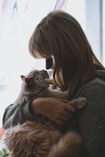 A person with medium-length hair gently holding a fluffy cat close, creating a tender moment. Soft light filters through a window in the background, illuminating the scene with a warm glow.