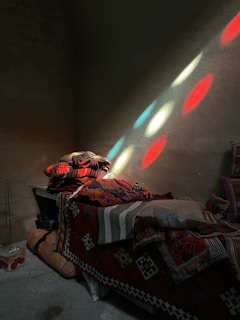 A dimly lit room with a stack of colorful patterned blankets and pillows on a bed. Sunlight filters through, casting vibrant red, green, and blue patches on the wall and the blankets. The overall setting has a rustic, cozy feel with an evident play of light and shadow.