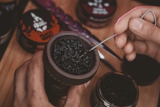 A hand is preparing a hookah bowl with dark tobacco, using a metal tool to adjust the mixture. Various containers of flavored tobacco are visible in the background on a wooden surface.
