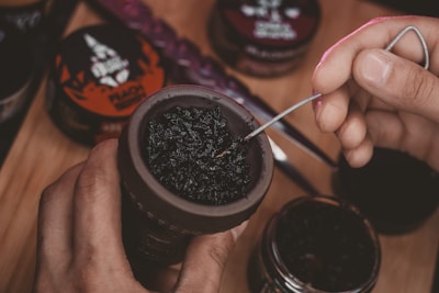 A hand is preparing a hookah bowl with dark tobacco, using a metal tool to adjust the mixture. Various containers of flavored tobacco are visible in the background on a wooden surface.