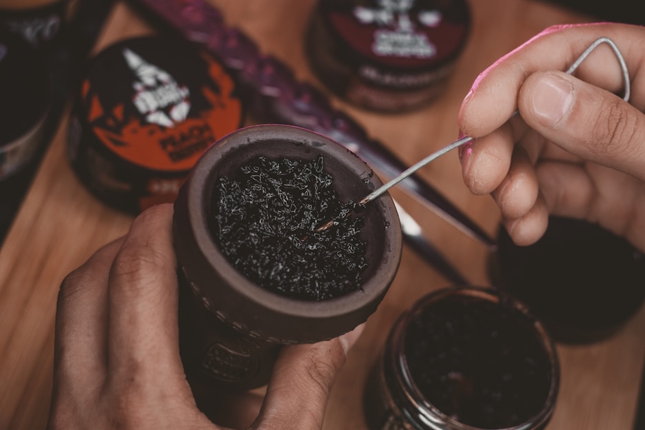 A hand is preparing a hookah bowl with dark tobacco, using a metal tool to adjust the mixture. Various containers of flavored tobacco are visible in the background on a wooden surface.