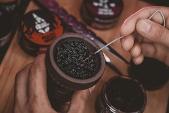 A hand is preparing a hookah bowl with dark tobacco, using a metal tool to adjust the mixture. Various containers of flavored tobacco are visible in the background on a wooden surface.